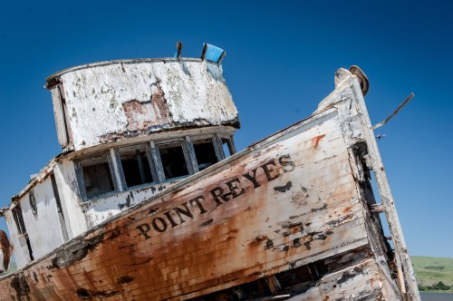 A weathered, abandoned boat named "Point Reyes" rests at an angle against a clear blue sky.