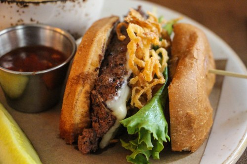 A close-up of a burger with lettuce, crispy onions, and melted cheese, served with a side of dipping sauce and a lemon wedge.