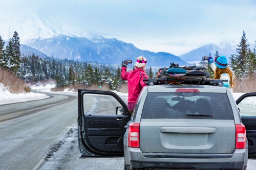 Two people in winter gear take photos from a parked car on a snowy road with mountains in the background.