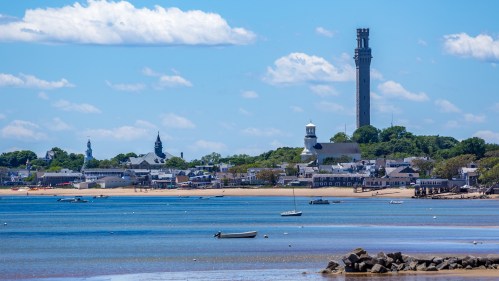 A coastal view of a town with boats in the water and a tall tower in the background under a blue sky with clouds.