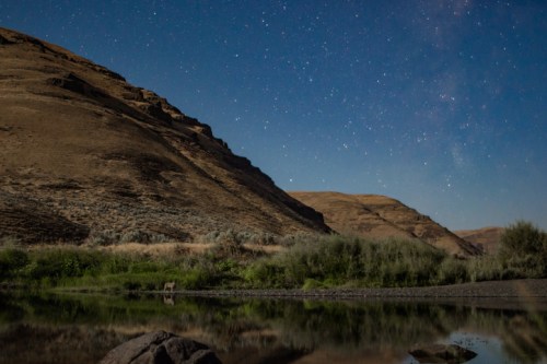 A serene landscape at night, featuring hills, a calm river, and a starry sky reflecting on the water's surface.