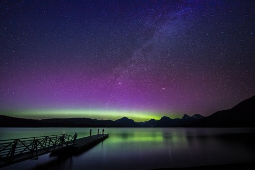 A serene lake at night with a colorful aurora and stars above, featuring a silhouetted pier and distant mountains.