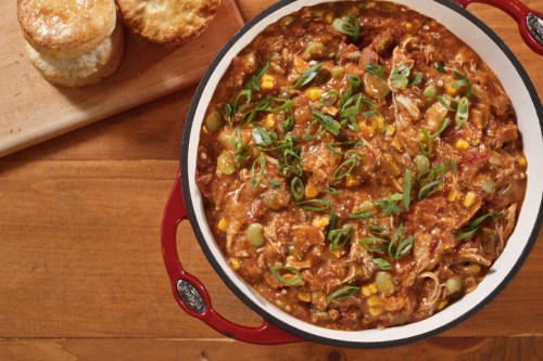A pot of hearty stew with green onions, corn, and shredded meat, alongside freshly baked biscuits on a wooden table.
