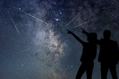 Silhouettes of two people pointing at a starry night sky filled with shooting stars and the Milky Way.