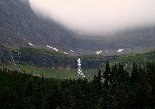 A misty mountain landscape with a waterfall cascading down rocky cliffs, surrounded by dense evergreen trees.