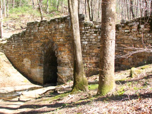 Stone bridge archway surrounded by trees in a wooded area, with a small stream nearby.
