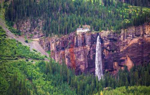 A stunning waterfall cascades down a rocky cliff, surrounded by lush green forests and a distant house perched above.