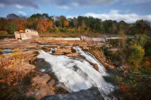 A scenic waterfall cascades over rocky terrain, surrounded by autumn foliage and a historic structure in the background.