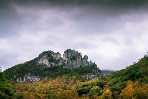 A rugged mountain peak rises above a forest of autumn-colored trees under a cloudy sky.