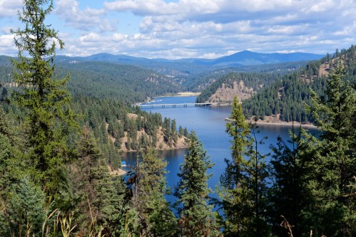 A scenic view of a lake surrounded by lush green forests and mountains under a partly cloudy sky.