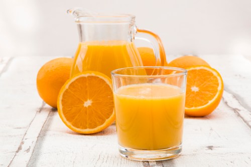 A glass of orange juice next to a pitcher of juice and fresh oranges on a wooden surface.