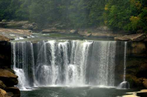 A serene waterfall cascading over rocks, surrounded by lush green trees and a calm river below.