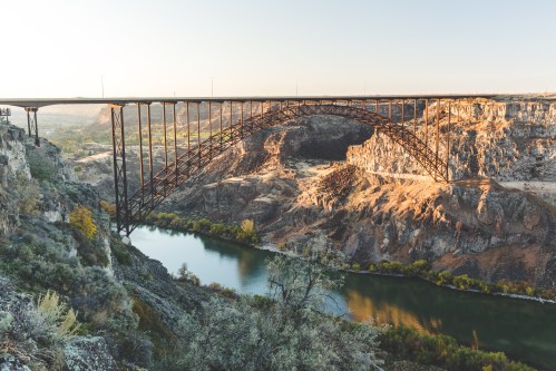 A large metal bridge spans a canyon, with a river below and rocky cliffs on either side, illuminated by soft sunlight.