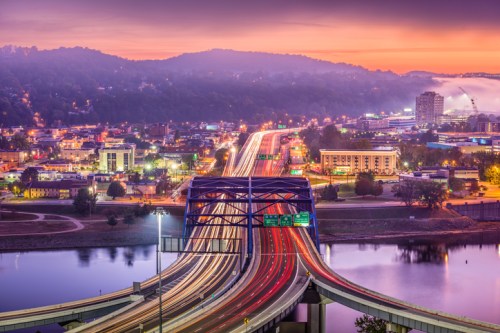 A vibrant cityscape at dawn, featuring a bridge over a river with light trails from vehicles and misty hills in the background.