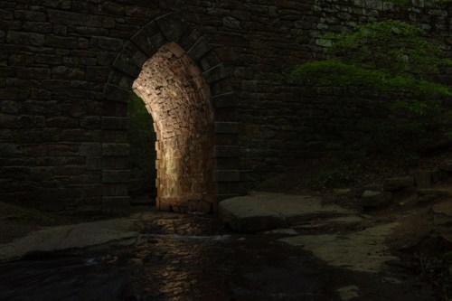 A stone archway illuminated from within, surrounded by dark foliage and a small stream in the foreground.