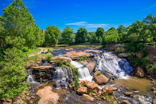 A scenic view of a waterfall surrounded by lush greenery and a clear blue sky. People walk along the riverbank.