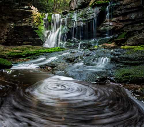 A serene waterfall cascades over rocks, surrounded by lush greenery and a tranquil pool with swirling water.