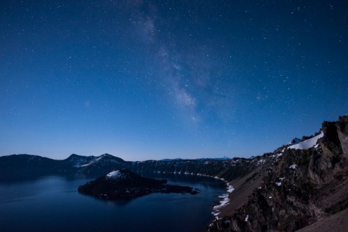 A serene night sky over a lake, with stars and the Milky Way visible above a snow-capped landscape.
