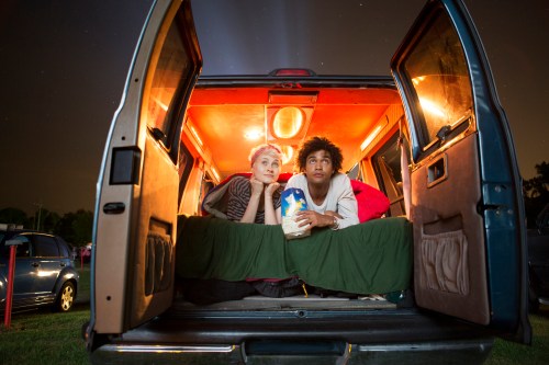 A young couple sits in a van at night, looking up at the stars, with a cozy blanket and popcorn between them.
