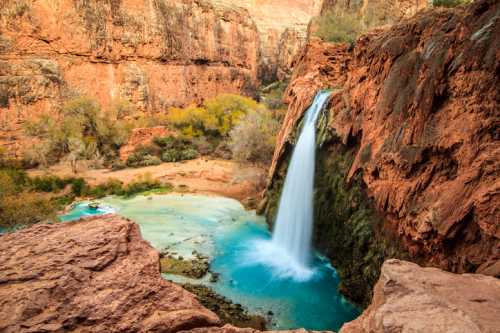 A stunning waterfall cascades into a turquoise pool, surrounded by red rock formations and lush greenery.