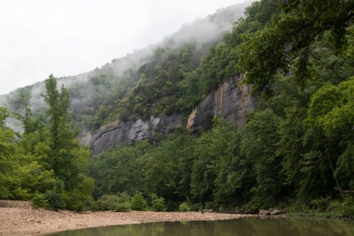 A misty mountain landscape with lush green trees and a calm river in the foreground.