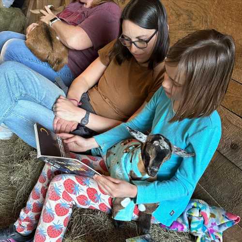 A woman and a girl sit on the ground, reading a book while holding a small goat, surrounded by others in a barn.