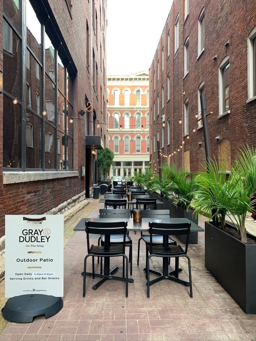 Outdoor patio with tables and chairs, surrounded by brick buildings and greenery, featuring a sign for Gray & Dudley.