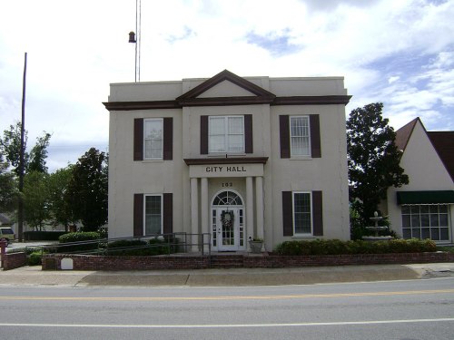 A two-story city hall building with a white facade, brown shutters, and a front staircase, surrounded by greenery.