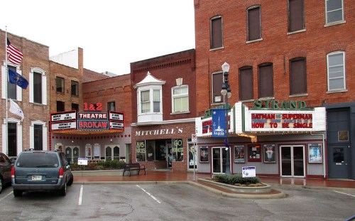 A street view of a theater district featuring marquee signs, brick buildings, and parked cars.