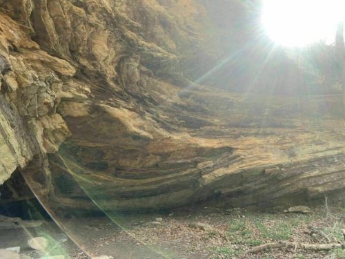 Sunlight streams through a rocky overhang, illuminating layered stone formations and a natural cave entrance.