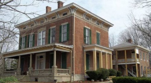 Historic brick house with green shutters, featuring a large porch and a side building, surrounded by trees.