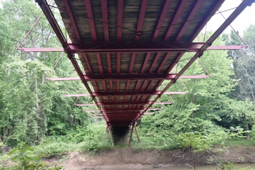 View from below a red metal bridge, surrounded by lush green trees and a calm riverbank.