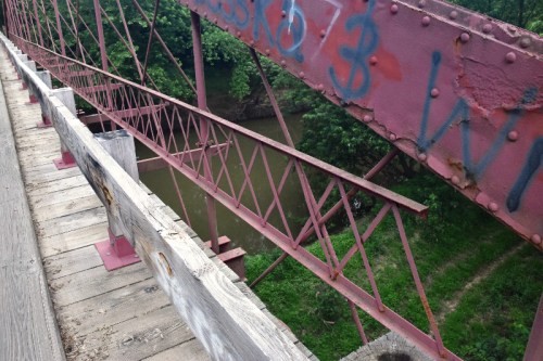 View from a bridge showing a rusted metal structure over a river, surrounded by greenery.