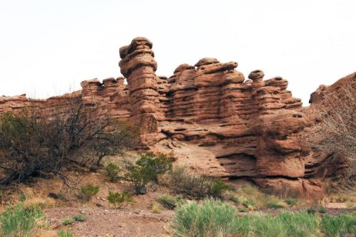 Rocky formations rise from the ground, surrounded by sparse vegetation and a clear sky in a desert landscape.