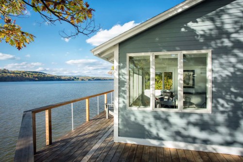 A modern gray house by the water, featuring large windows and a wooden deck overlooking a scenic river landscape.