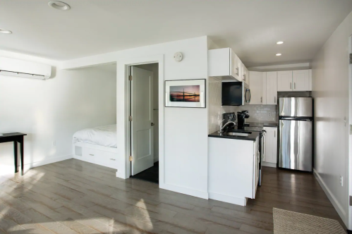 Bright, modern studio apartment with a kitchenette, dining table, and a bed visible through an open door.