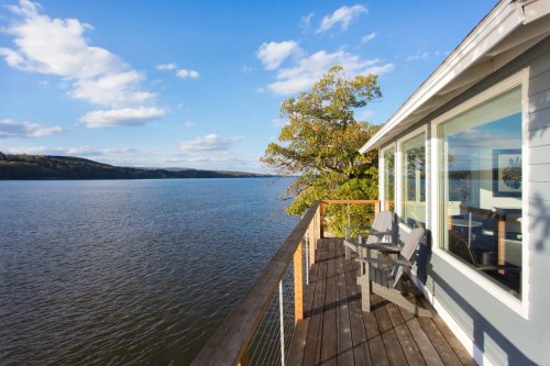 A serene lakeside view from a deck with two chairs, surrounded by trees and a clear blue sky.