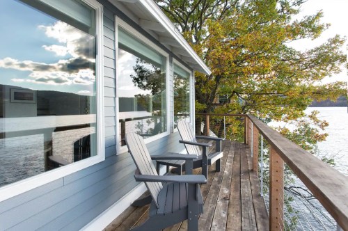A lakeside deck with two chairs, reflecting windows, and trees in the background under a cloudy sky.