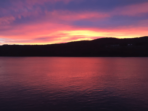 A serene lake at sunset, reflecting vibrant pink and purple hues against a dark silhouette of hills.