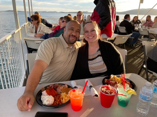 A couple smiles at the camera while enjoying a meal on a ferry, with water and other passengers in the background.
