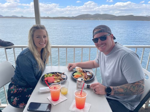 A couple enjoys a meal with drinks at a waterfront table, surrounded by scenic views of water and mountains.