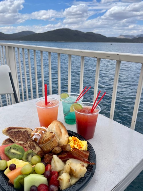 A colorful brunch spread with fruit, pastries, and three vibrant drinks on a table by a lake with mountains in the background.