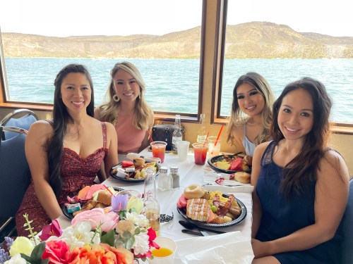 Four women smiling at a table with food and drinks, with a scenic view of water and mountains in the background.
