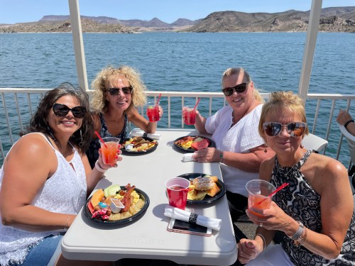 Four women enjoying a meal and drinks on a boat, with a scenic lake and mountains in the background.