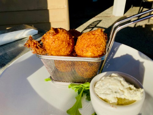 A small basket of golden-brown fried balls served with a side of creamy dip on a white plate.