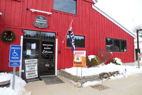 A red barn-style building with signs for a bakery and mercantile, surrounded by snow and parking spaces.