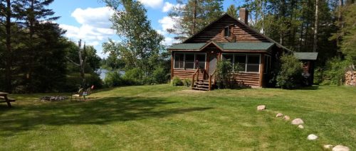 A rustic log cabin surrounded by trees, with a lawn leading to a lake under a blue sky with fluffy clouds.