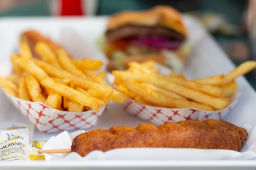 A tray with golden French fries, a burger with toppings, and a corn dog, accompanied by mustard packets.