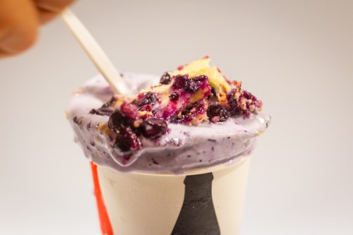 A close-up of a cup of purple ice cream topped with berries and cake pieces, with a spoon inserted.