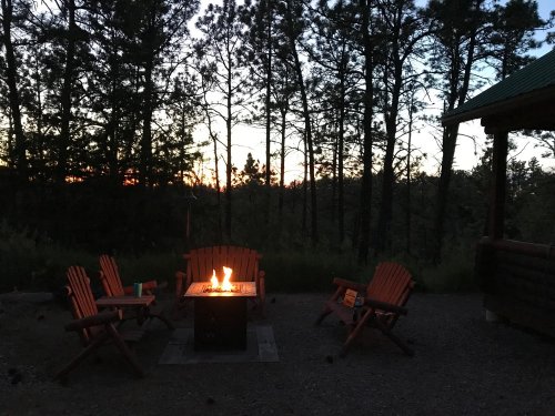 A cozy fire pit surrounded by wooden chairs, set against a backdrop of trees and a sunset sky.